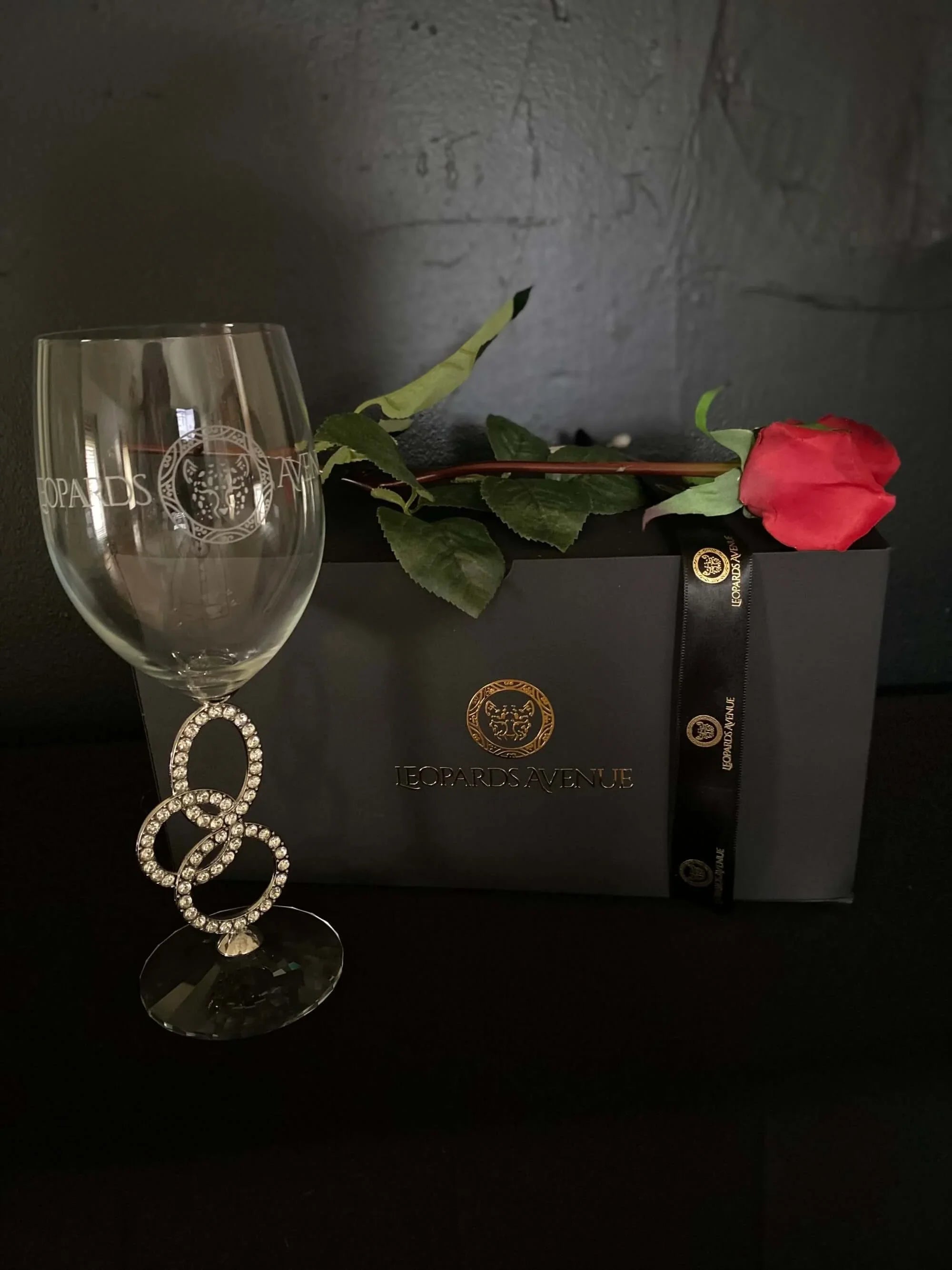 Leopards Avenue wine glass with jeweled stem, black gift box, and red rose on black background