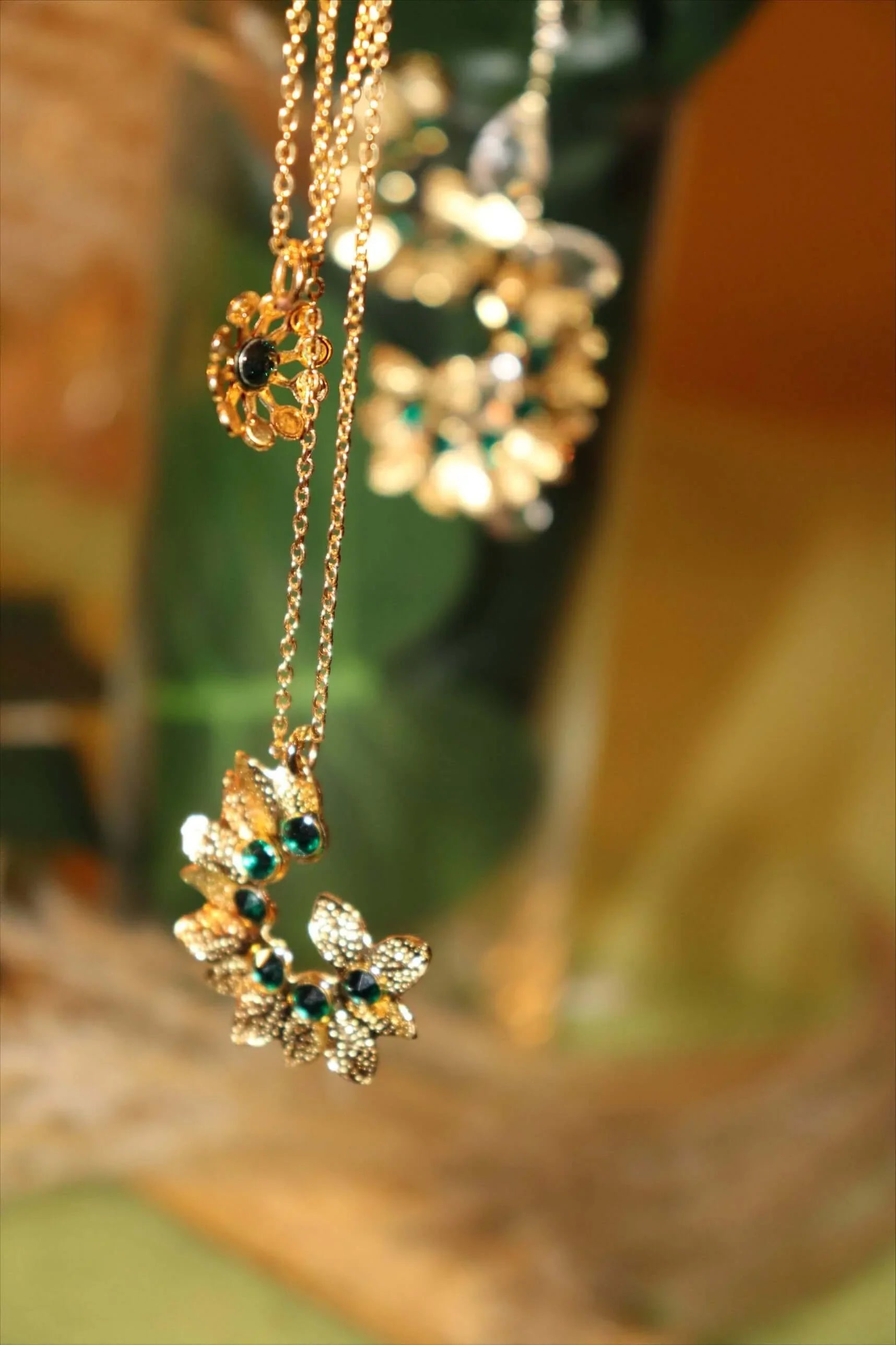 Gold flower pendant necklace with green gemstones on delicate chain, blurred natural background.