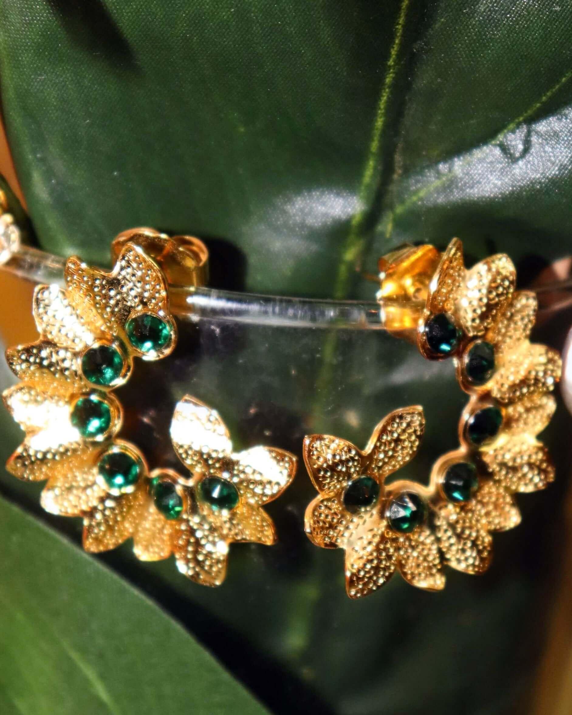 Gold floral hoop earrings with green gemstones displayed on a green leaf background